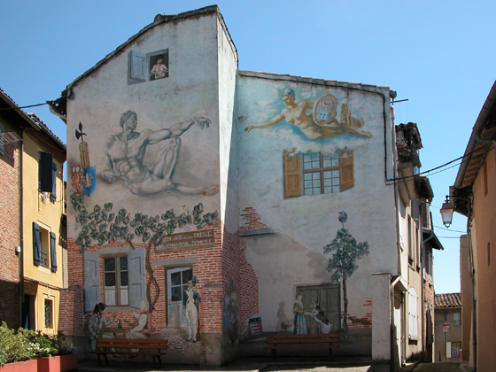 Façade nue d'une maison décorée de peintures en trompe l'oeil. Dans la partie supérieure : Une déesse Grecque, un dieu Grec, une femme à une fenêtre. Dans la partie inférieure, un soldat républicain monte la garde devant la porte ouverte d'une auberge, un couple se désaltère, assis autour d'une table placée à l'extérieur, une femme joue avec un chien. 